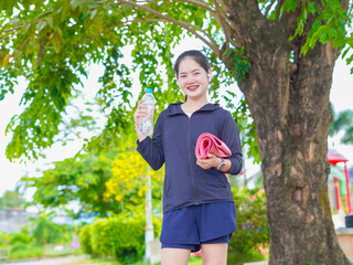 Asian fitness woman holding water bottle and yoga mat standing in a park, a natural environment. Self-care and health. Southeast Asian wellness lifestyle work-life balance, mental wellbeing. Workout 