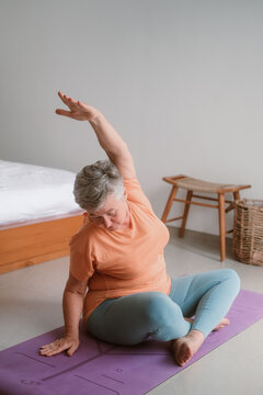 A senior woman with gray hair sits in a lotus position on a yoga mat in her room. She does exercises and gymnastics for the elderly and joint support. Health improvement in retirement.