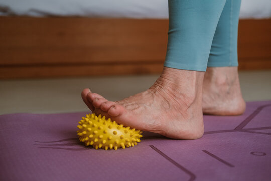 Close-up of a senior woman foot standing on a mat and massaging her foot with a rubber ball with spikes. Wellness massage for the rehabilitation of the elderly. Body care in retirement.