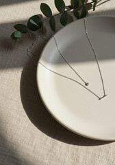 Minimalist silver necklace displayed on a ceramic plate with soft beige fabric. Clean aesthetic and elegant flat lay composition.