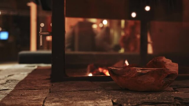 A modern fireplace with flickering flames is surrounded by warm lighting. A wooden bowl on a stone surface adds a rustic touch to the inviting scene.