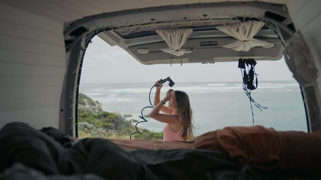 Woman uses a handheld shower outside her van, gazing at the ocean waves under a clear sky