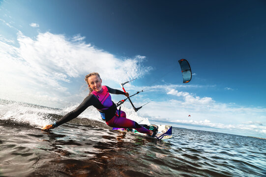 Kitesurfing girl in sexy wetsuit with kite in sky on board in blue sea riding on waves with water splash. Recreational activity, water sports, action, hobby and fun in summer. Kiteboarding sport