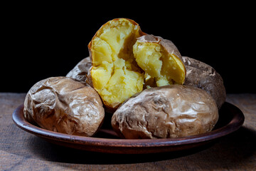 Baked potatoes whole in their skins on a clay plate, closeup. Simple Ukrainian food on black background. Jacket potato for eat