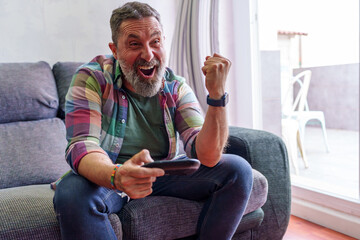 Bearded gamer sitting relaxed on sofa, holding controller and celebrating triumphant moment during...