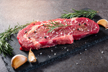 Seasoned raw beef steak with sea salt, cracked pepper, rosemary sprigs, and garlic cloves on black slate. High-end food styling for grilling, recipe visuals, or meat preparation advertising