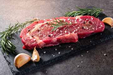 Seasoned raw beef steak with sea salt, cracked pepper, rosemary sprigs, and garlic cloves on black slate. High-end food styling for grilling, recipe visuals, or meat preparation advertising