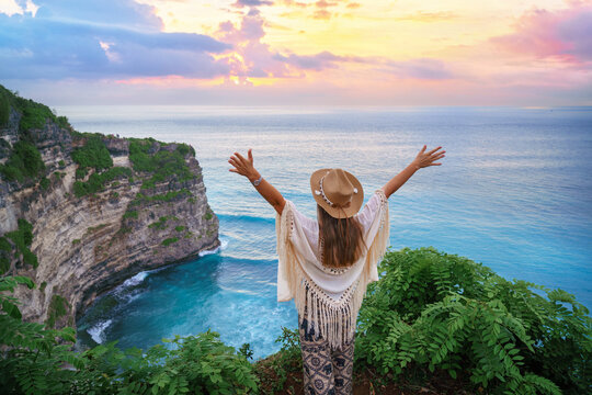 Happy free traveler girl wearing hat with open arms enjoys turquoise stunning ocean view on cliff during traveling at beautiful destination on Bali island, Indonesia