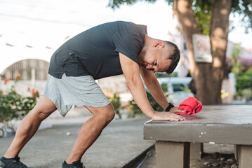 Middle-aged Southeast Asian man leans on park bench after jogging, appearing dizzy or overheated,...