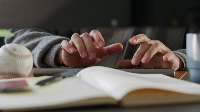 Cropped shot of hands of unrecognizable person in casualwear sitting at computer desk and typing on pc keyboard with opened copybook in blurred foreground