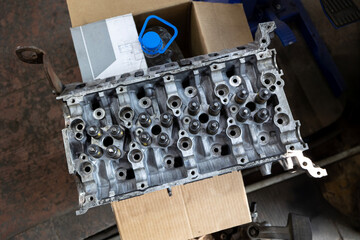 An overhead close-up shows a cylinder head with valves resting inside a cardboard box in a workshop...