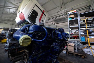 Naklejka premium A disassembled diesel truck engine stands on wooden supports in a repair shop or warehouse, with a truck cab tilted in the background