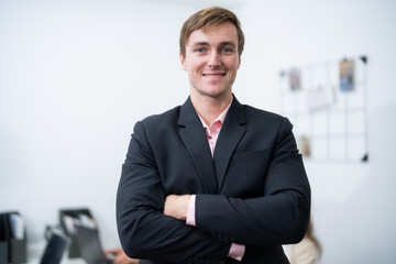 Fototapeta premium Confident young man poses in a business office setting with arms crossed and a smile during working hours