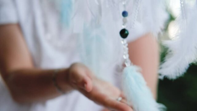 A dreamcatcher with white feathers and ribbons sways in the wind. Women's hands touch and adjust the ribbons.