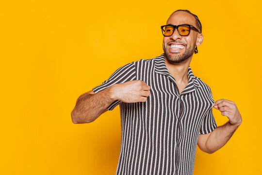 Portrait of a fashionable man wearing yellow sunglasses and a striped shirt, confidently pointing at himself against a vibrant yellow background.