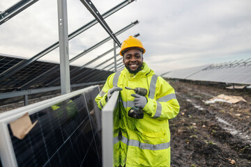 Portrait of cheerful black repairman standing at solar farm next to solar panels and holding drill...