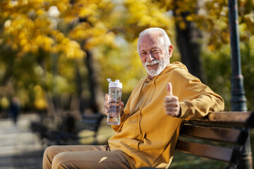 Portrait of senior sportsman sitting on a bench in park with bottle of water in hands and giving...