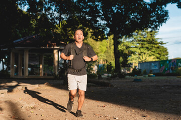 Middle aged Southeast Asian man runs through park wearing weighted backpack, captured mid-stride during an intense rucking workout.