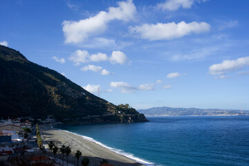 Obraz premium A sweeping view of a pebbled beach along the coastline of Scilla with palm trees, azure sea, and distant mountain ranges beneath a vibrant blue sky with scattered clouds