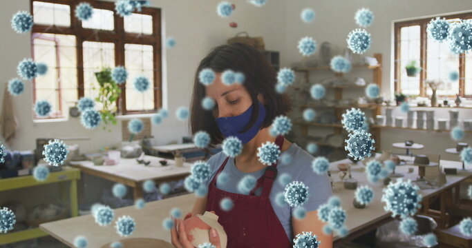Composite image of covid-19 cells floating against female potter wearing a face mask holding clay - Powered by Adobe