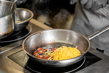 A chef in black gloves adds cooked spaghetti to a skillet with sizzling bacon bits and steam rising, capturing the essence of culinary preparation in a busy kitchen.