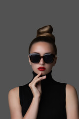 Young stylish woman in black sunglasses and red lips makeup showcases elegance. Her polished blonde bun and black turtleneck showcase eye wear fashion look against a dark gray background.