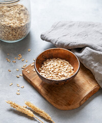 Oat flakes in bowl on wooden board with napkin, wheat and glass jar on grey textured background.