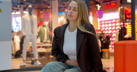 Obraz premium Woman with stomach pain sits on bench in shopping mall, eyes closed.