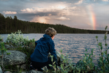 A boy sits crouched against rocks by the water, looks at the vibrant rainbow and sky as evening falls over the tranquil landscape of Dalarna, Sweden