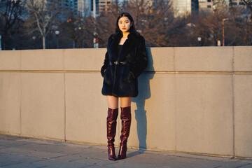 A woman leans against a wall wearing a black dress and knee-high boots