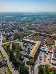 Aerial view of a modern building complex with solar panels, surrounded by residential houses and green areas, located near sandy dunes and walking trails under a clear blue sky.