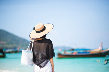 The back of one Asian woman wearing a shirt and a hat with holding bag walks on a sunny beach with blue sea and boats in the background. Concept of holidays and vacations. Picture looking alone or sad