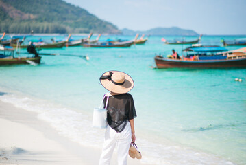 Back of Asian woman with holding a hat on a sunny beach with sea and boats in the background. Concept of holidays and travel. Picture looking alone or sad. She looking forward.