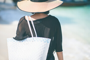 The back of one Asian woman wearing a shirt and a hat with a holding the bag with blue sea and boats in the background. Concept of holidays and vacations. Picture looking alone.