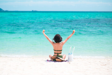 Back of Asian women look happy wearing green bikini outstretched arm sitting on the beach with blue sea and sky background in the daytime. Concept of happy holidays and travel. Copy space for text.