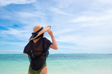 Back of Asian woman wearing See-through and green bikini holding a hat on a sunny beach with sea and sky background in the daytime. Concept of long weekend holidays and travel. Copy space for text.
