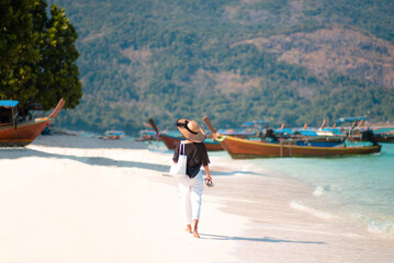 Long shot back of Asian woman wearing shirt and a hat with holding bag walks on a sunny beach with blue sea and boats in the background. Concept of holidays and travel. Picture looking alone or sad