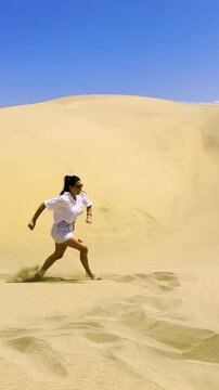 Slowmo of Joyful Woman Jumping in Desert Sand Dunes Under Bright Blue Sky. Happy traveler jumps barefoot in golden sand dunes with arms raised, capturing freedom and joy in a vibrant desert landscape
