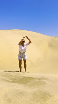 Slowmo of Joyful Woman Jumping in Desert Sand Dunes Under Bright Blue Sky. Happy traveler jumps barefoot in golden sand dunes with arms raised, capturing freedom and joy in a vibrant desert landscape
