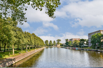 Fototapeta premium A picturesque view of a serene river gently flowing alongside a line of green trees, under a bright, partly cloudy sky. A peaceful escape into nature.