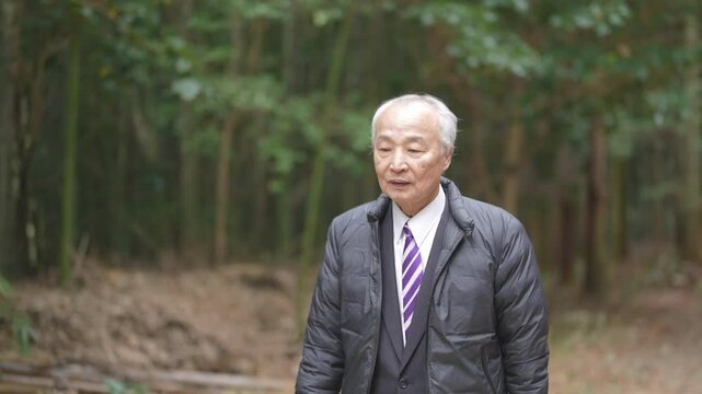 A winter park in Japan. A forest with many trees growing. A 78-year-old Japanese man wearing a down jacket and suit stands. He's walking.