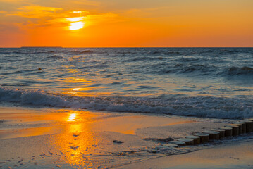 Obraz premium Sunset over the Baltic Sea. The bright orange sun gradually sinks below the horizon, illuminating the clouds and giving the water a warm golden-orange hue. 
