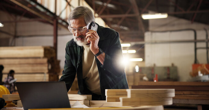 Phone call, laptop and man in carpentry workshop for furniture, manufacturing and lumber renovation. Construction, factory and carpenter on smartphone for online orders, inventory and timber stock