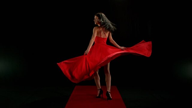 Stunning Black-Haired Woman in Ruby Red Dress and High Heels Gracefully Posing Against a Black Background, Captured in Super Slow Motion for a Dramatic and Elegant Fashion Cinematic Effect.