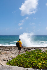 Back View of a Man with Yellow T-Shirt, Baseball Cap and Black Backpack Standing in Front of the Caribbean Ocean Watching Water Shooting Out from a Blow Hole