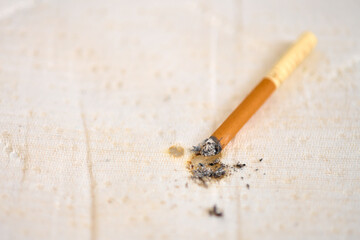 Single burned cigarette rests on textured beige fabric surface. Soft lighting highlights ash remnants and brown filter. Close-up perspective emphasizes cigarettes details