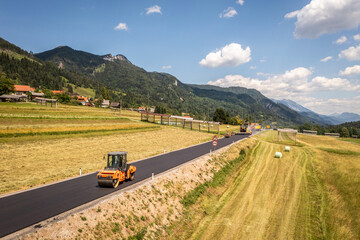 Fototapeta premium Drone view of compact roller smoothing fresh asphalt on new countryside road during final paving stage