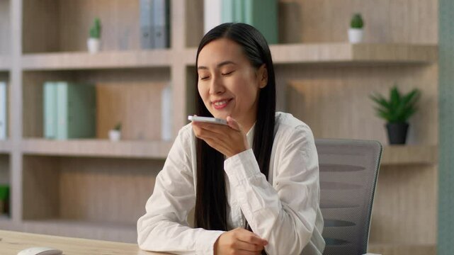 Joyful Chinese businesswoman worker Korean Japanese business woman female manager using mobile phone online app call talking smartphone speakerphone recording voice recognition message send SMS indoor