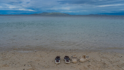 Two pairs of flip-flops on a sandy beach by the turquoise ocean. Women's burgundy with artificial flowers, men's ivory with buckles. Shells, coral fragments on the sand. Mountains on the horizon.  