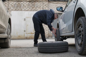 A man in a black jacket and trousers changes winter tires to summer tires on a car, a wheel lies next to it